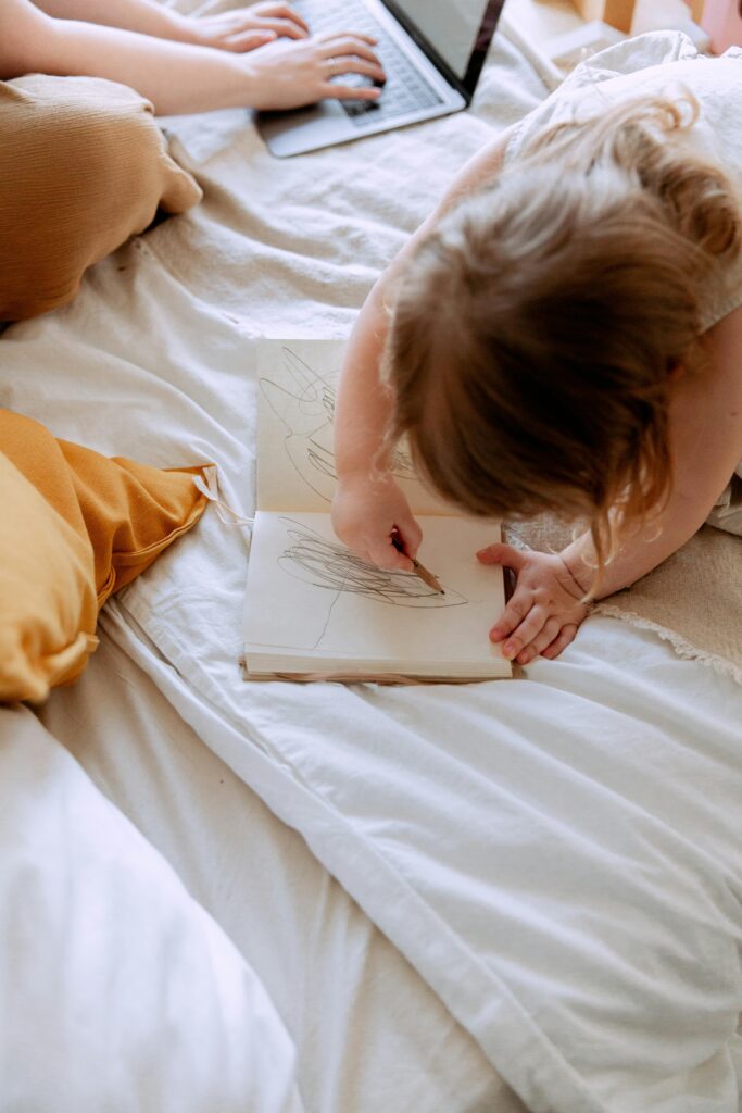 A woman in bed working on her laptop attempting to balance work and parenting while her daughter draws on a notebook beside her.